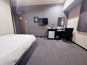 A hotel room with a bed, a desk with a computer, a small fridge, and a TV mounted on the wall.
