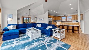A spacious living room with a blue sectional sofa, white coffee tables, and a starfish-patterned rug. A ceiling fan and recessed lighting are visible. - 7135-Our Blue Haven - Waves Retreat | Pool | 300 Yards to Beach (Waves)