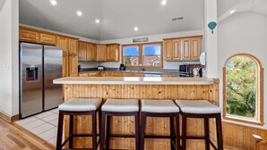 A kitchen with wooden cabinets, stainless steel appliances, a large island with bar stools, and a view of the outdoors through a window. - 7135-Our Blue Haven - Waves Retreat | Pool | 300 Yards to Beach (Waves)