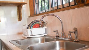 A kitchen with two stainless steel sinks, a dish rack with plates and utensils, and a window with blinds. - Van Villa Holidays (Negril)