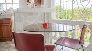 A kitchen with a glass table and red chairs, a window with a view of greenery, and a white tiled backsplash. - Van Villa Holidays (Negril)