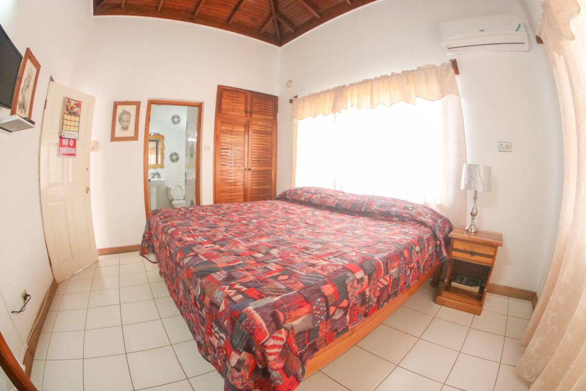 Quarto com cama, mesa de cabeceira, janela com cortinas, porta e ar-condicionado fixado na parede.