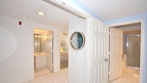 Un pasillo con pisos de baldosas blancas, puertas blancas y un baño con lavamanos y espejo.