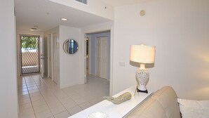 Un salón con un sofá blanco, una mesa de centro blanca con objetos decorativos, una lámpara y una puerta que da a otra habitación.