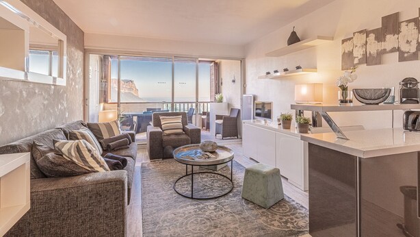 A modern living room with a sofa, armchairs, and a coffee table. There is a kitchen area with a countertop and appliances. Large windows offer a view of the outdoors. - L'épuisette à Cassis by Winkyhouse (Cassis)