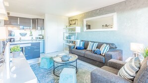 A modern living room with a grey sofa, a coffee table, and a blue rug. There is a kitchen area with cabinets and a microwave. - L'épuisette à Cassis by Winkyhouse (Cassis)