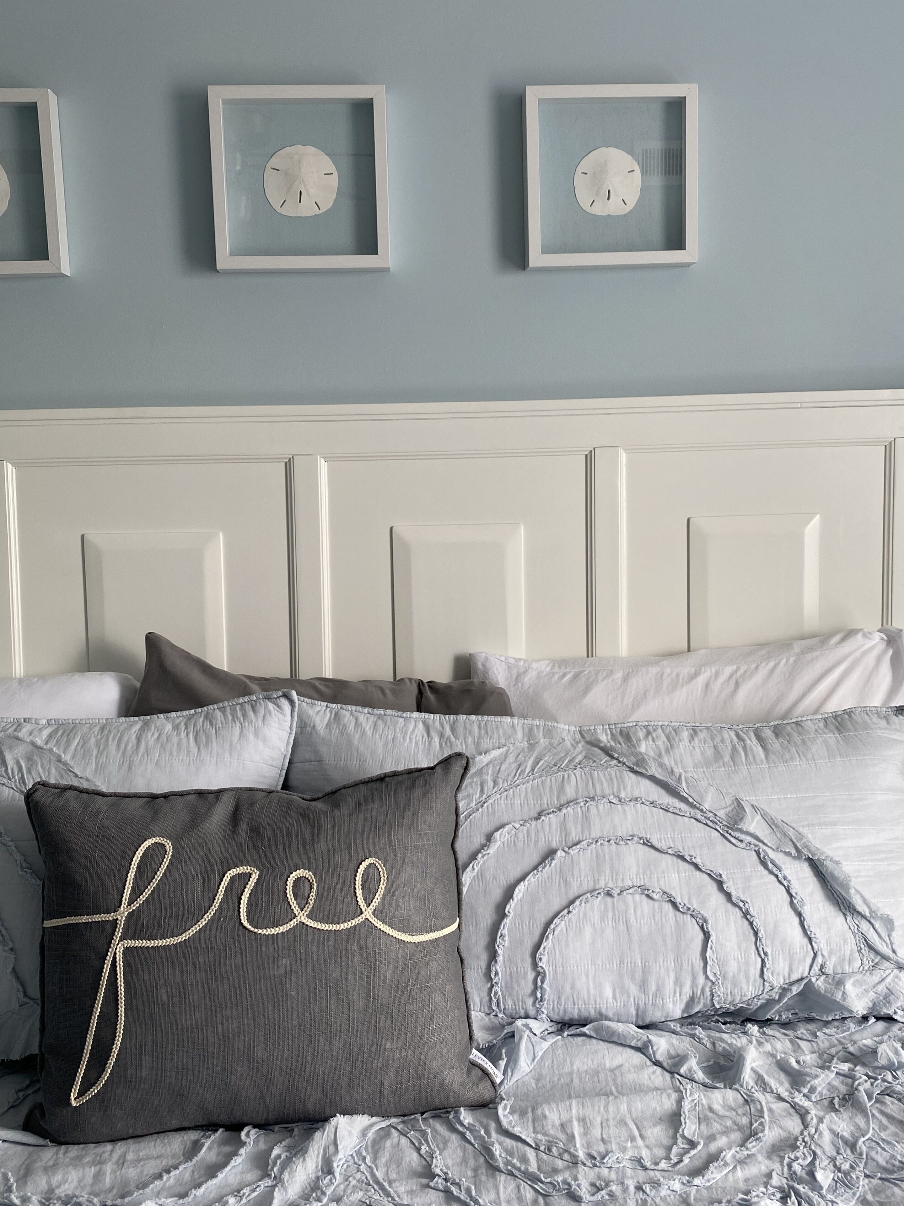 A bedroom with a white headboard, grey bedding, and a pillow with the word "free" embroidered on it.