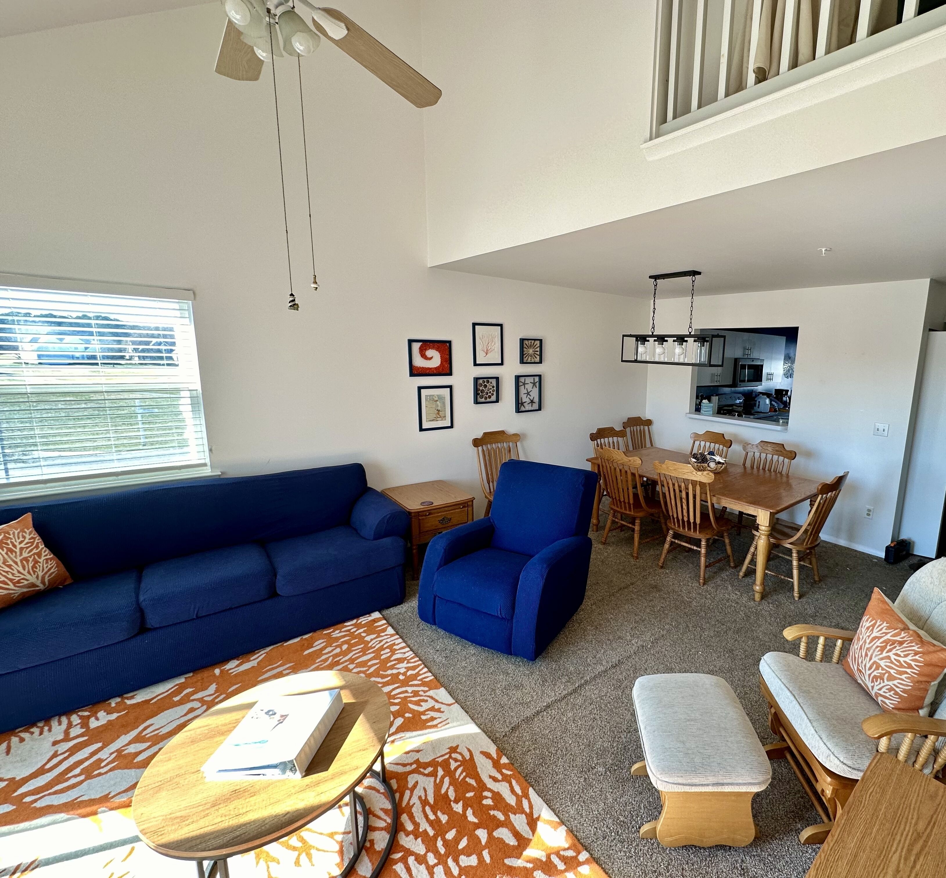 A living room with a blue sofa, a blue armchair, and a wooden coffee table. There is a dining area with a wooden table and chairs, and a kitchen in the background.