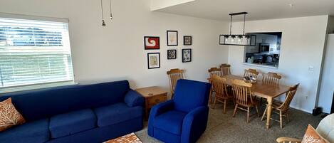 A living room with a blue sofa, a blue armchair, and a wooden coffee table. There is a dining area with a wooden table and chairs, and a kitchen in the background.