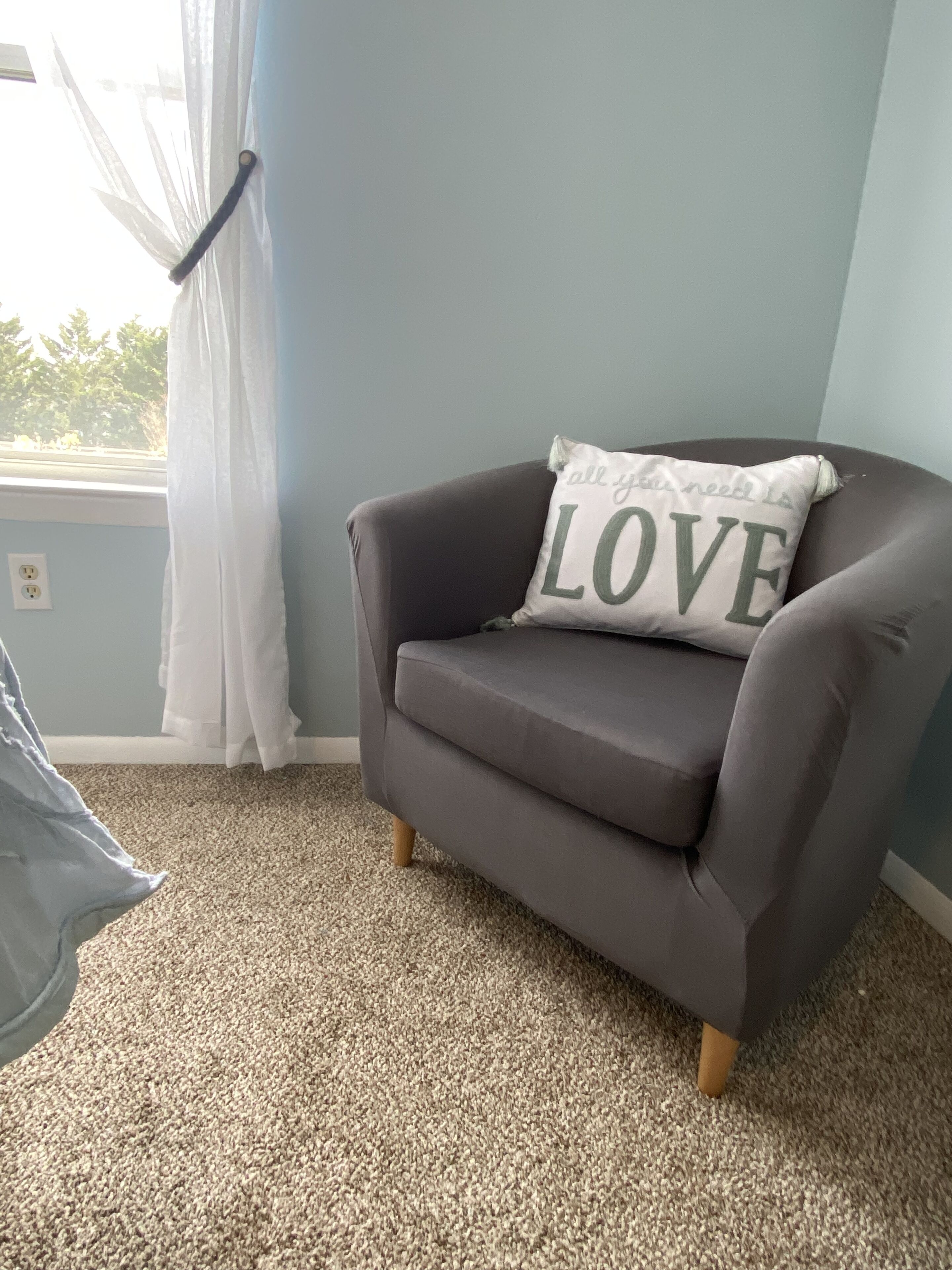 A grey armchair with a pillow that reads "all you need is LOVE" against a light blue wall, a window with sheer white curtains, and a beige carpet.