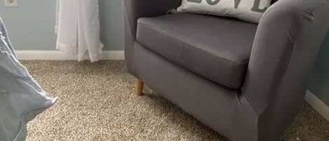 A grey armchair with a pillow that reads "all you need is LOVE" against a light blue wall, a window with sheer white curtains, and a beige carpet.