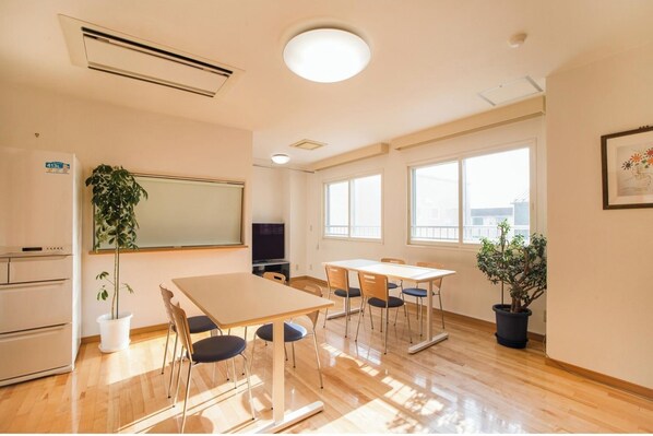 Une pièce dotée d’un grand tableau blanc, d’un téléviseur à écran plat, de tables et de chaises en bois, de plantes en pot et de grandes fenêtres.