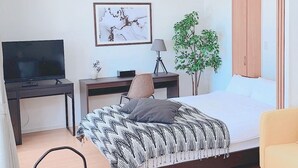 Ein Schlafzimmer mit einem Bett, einem Schreibtisch, einem Fernseher, einer Lampe und einer Pflanze.