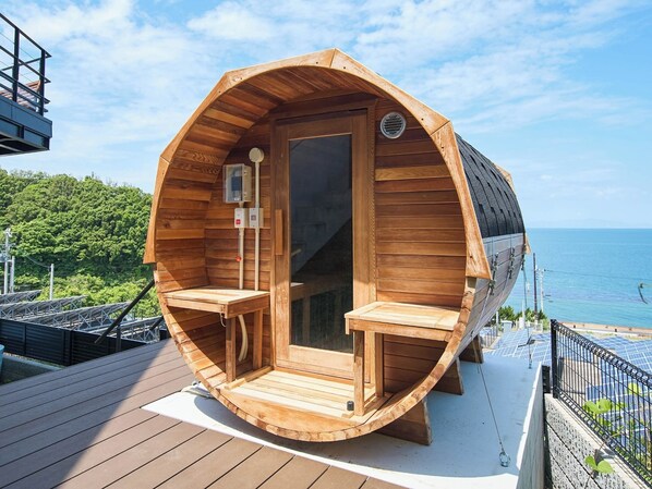 Una estructura cilíndrica de madera con puerta y ventanas, una pequeña mesa y un banco, ubicada en una terraza con paneles solares y vista al mar.