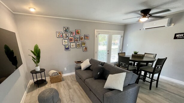 A modern living room with a grey sofa, round ottoman, and a dining table with chairs. There are wall-mounted air conditioning units and a ceiling fan.
