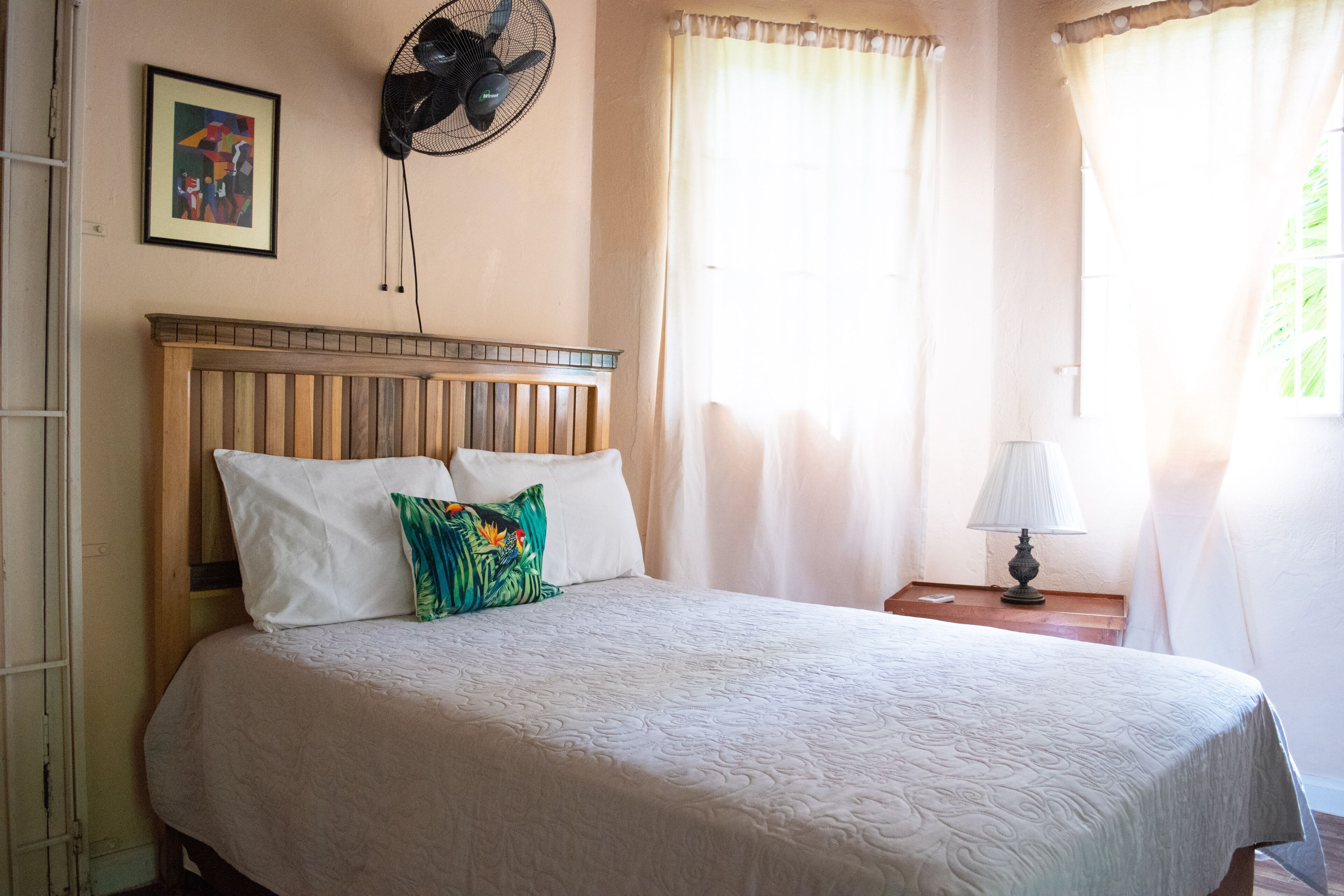 A neatly made bed with a wooden headboard, a bedside table with a lamp, and a wall-mounted fan.