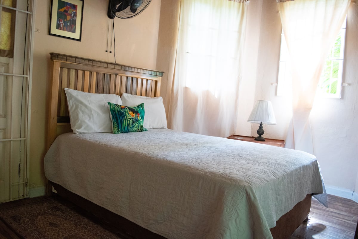 A neatly made bed with a wooden headboard, a bedside lamp, and a window with sheer curtains.