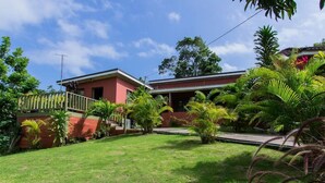 A two-story building with a red exterior and a balcony. The property has a well-maintained lawn and various tropical plants.