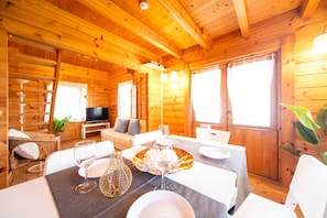Interior de uma cabana de madeira com uma mesa de jantar posta para dois, um sofá e uma televisão ao fundo.