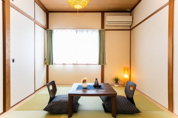 Une pièce de style japonais traditionnel, avec un sol recouvert de tatamis, une petite table basse avec des coussins, un luminaire suspendu et une fenêtre avec des rideaux.