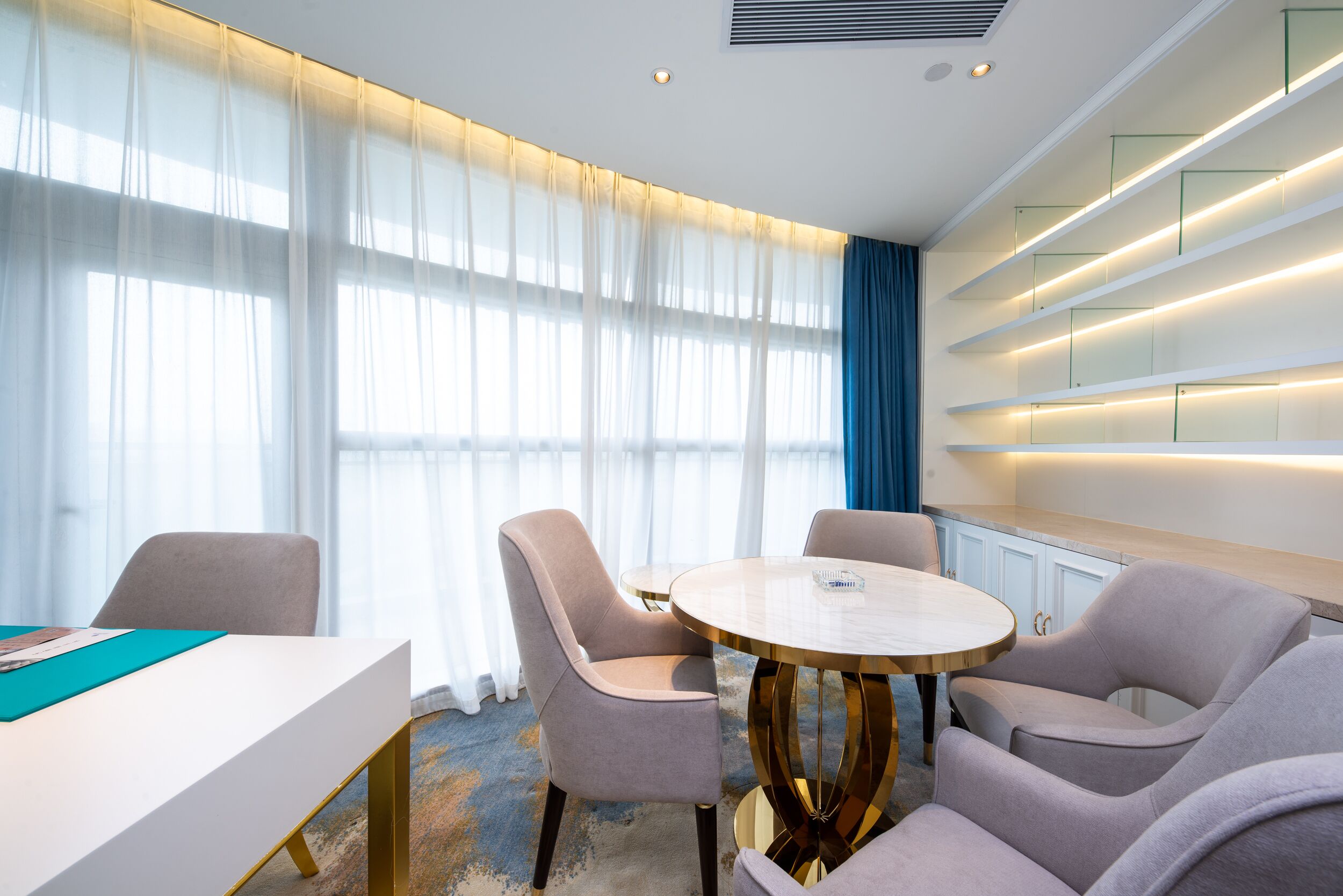 Een moderne hotelkamer met een ronde tafel, twee stoelen, een wit bureau en een groot raam met dunne gordijnen.