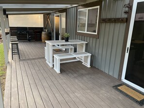 A covered patio with a wooden deck, white tables and benches, a grill, and a sliding glass door.