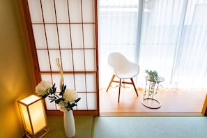 Une pièce de style japonais traditionnel, avec un sol recouvert de tatamis, un paravent shoji, une chaise blanche, une petite table ornée d’une plante et une lanterne allumée.
