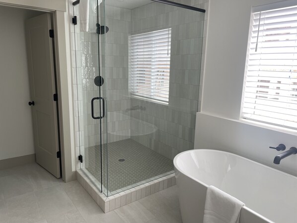 A modern bathroom with a glass-enclosed shower, a freestanding bathtub, and a window with blinds.