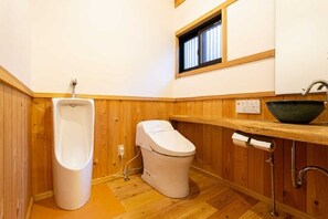 A small, compact bathroom with a wooden floor, a white toilet, a urinal, a sink with a green basin, and a window above the sink.