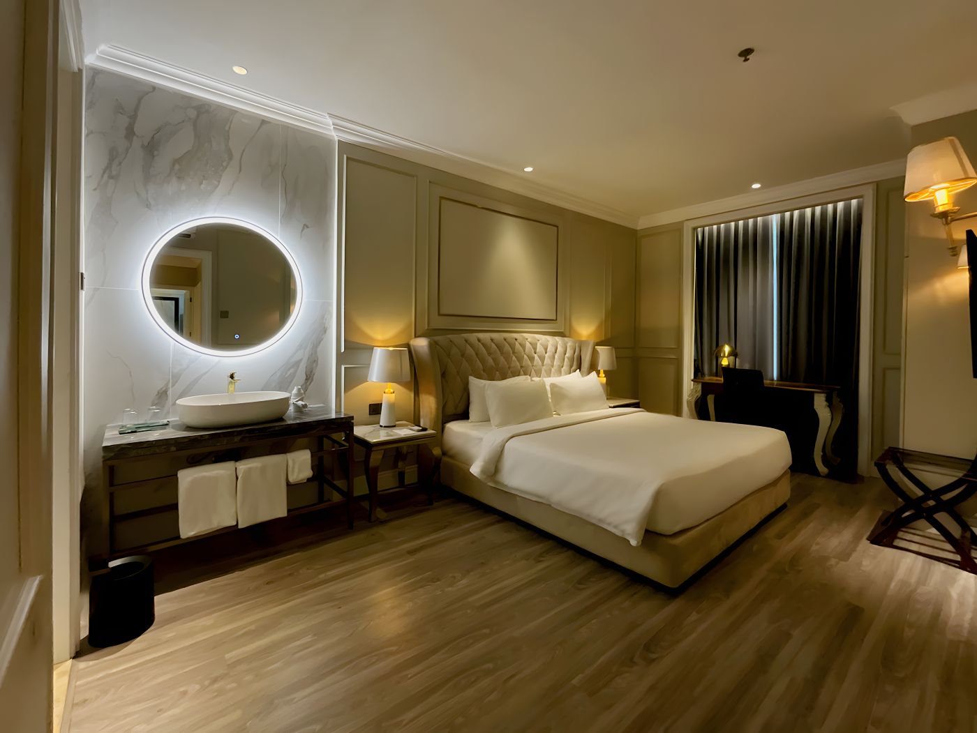 Une chambre d’hôtel avec un grand lit, un miroir rond, une coiffeuse avec un lavabo et une table de chevet avec des lampes.