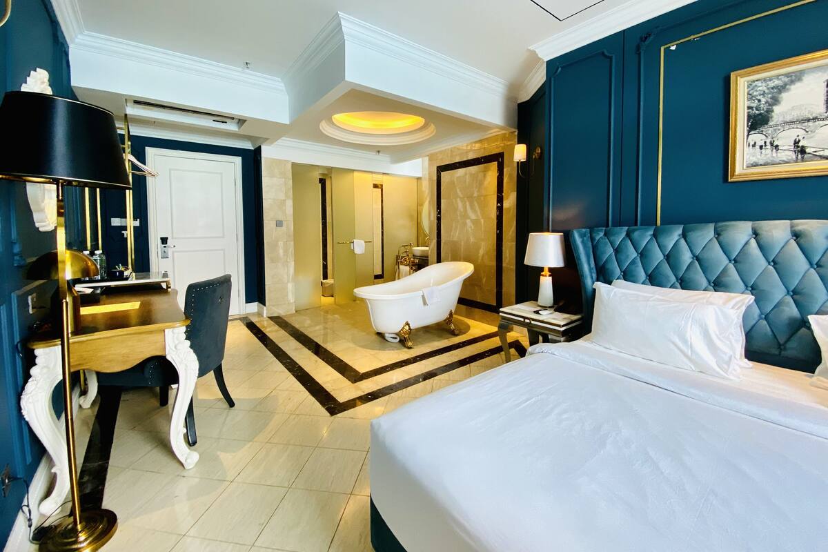 A hotel room with a large bed, a sitting area with a chair, a desk, a vanity, and a bathtub.