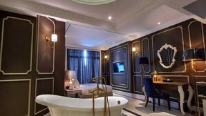 A luxurious bathroom with a freestanding bathtub, a vanity with a mirror, and a seating area with a chair and table. - Grand Eska Hotel & Suite  (Batam)