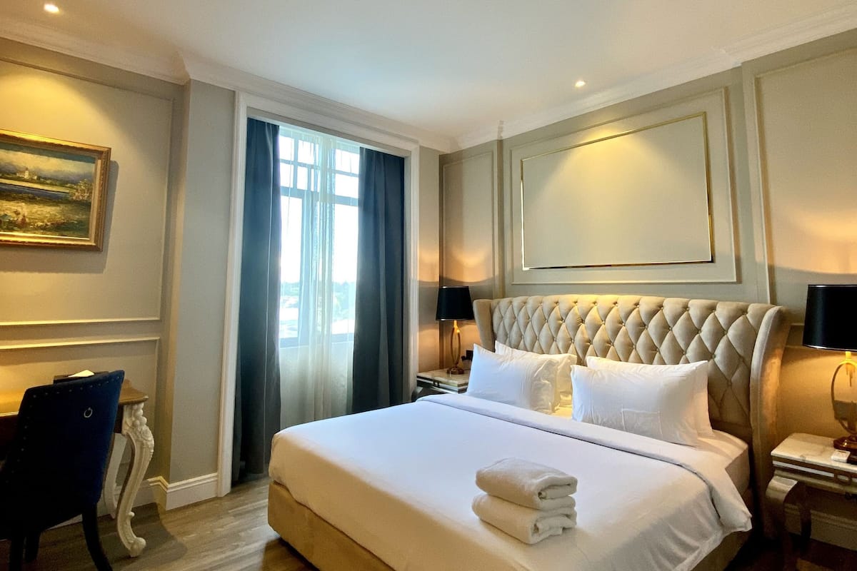 A large bed with a tufted headboard, a bedside table with a lamp, a chair, and a painting on the wall.