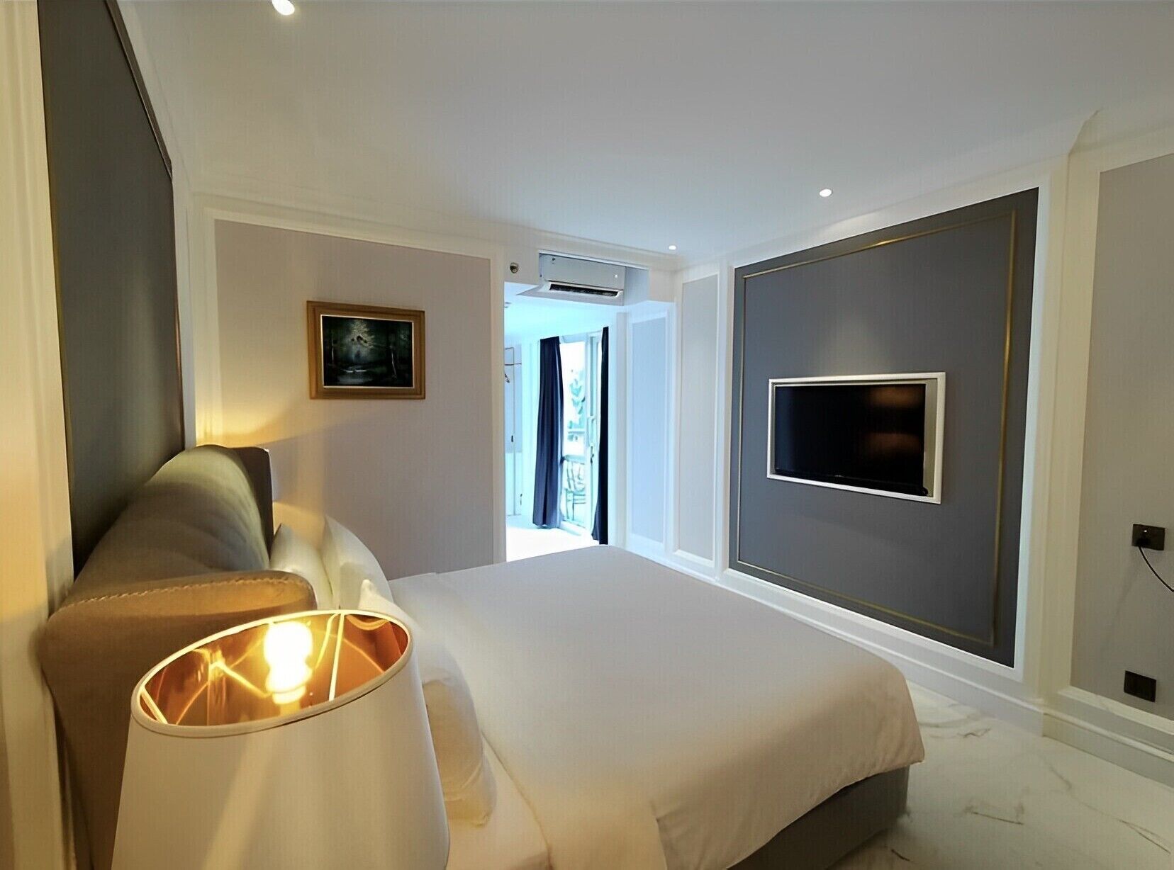 A modern hotel room with a large bed, a bedside lamp, a flat-screen TV mounted on the wall, and a painting on the wall.