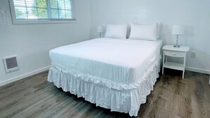 A bedroom with a large bed, white bedding, a nightstand with a lamp, and a window with blinds.
