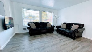 A spacious living room with two black leather sofas, a floor lamp, and a large window with blinds.