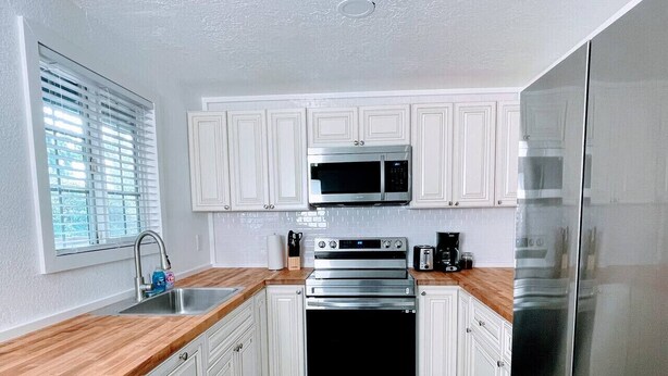 A kitchen with white cabinets, a stainless steel oven, a microwave, a refrigerator, and a wooden countertop.