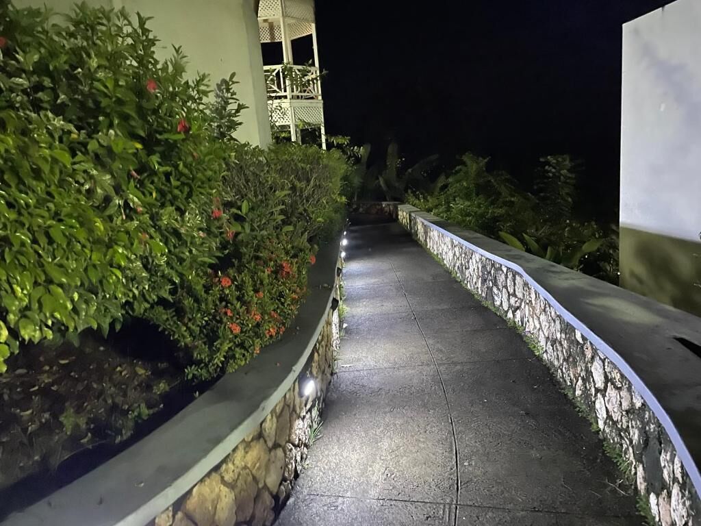 A nighttime pathway flanked by stone walls and lush greenery, leading to a building with a balcony.
