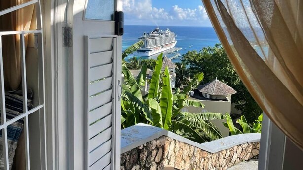 A balcony with a view of a cruise ship on the ocean, surrounded by greenery and a stone wall. - Humming Bird Holiday Retreat (Ocho Rios)