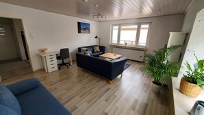 Ein modernes Wohnzimmer mit einem blauen Sofa, einem Holztisch und einem Schreibtisch mit Stuhl. Ein großes Fenster bietet Blick ins Freie, und eine Topfpflanze steht rechts.