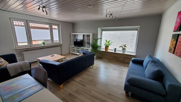 Ein Wohnzimmer mit blauem Sofa, einem Holztisch, einem Fernseher auf einem weißen Schrank und einem großen Fenster mit Blick.