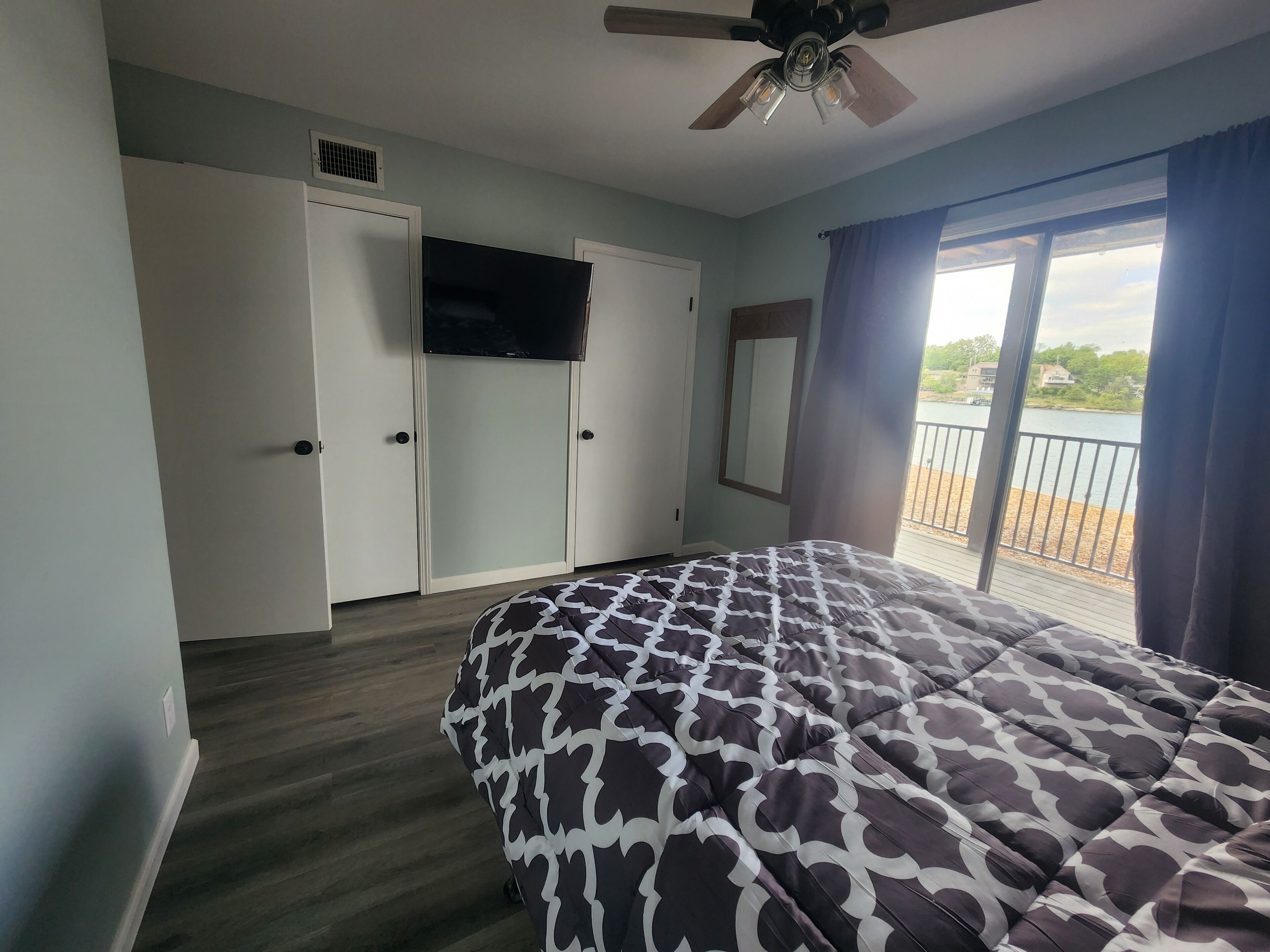 A bedroom with a bed, a ceiling fan, a TV, and a view of a lake through a window.