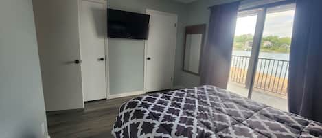 A bedroom with a bed, a ceiling fan, a TV, and a view of a lake through a window.