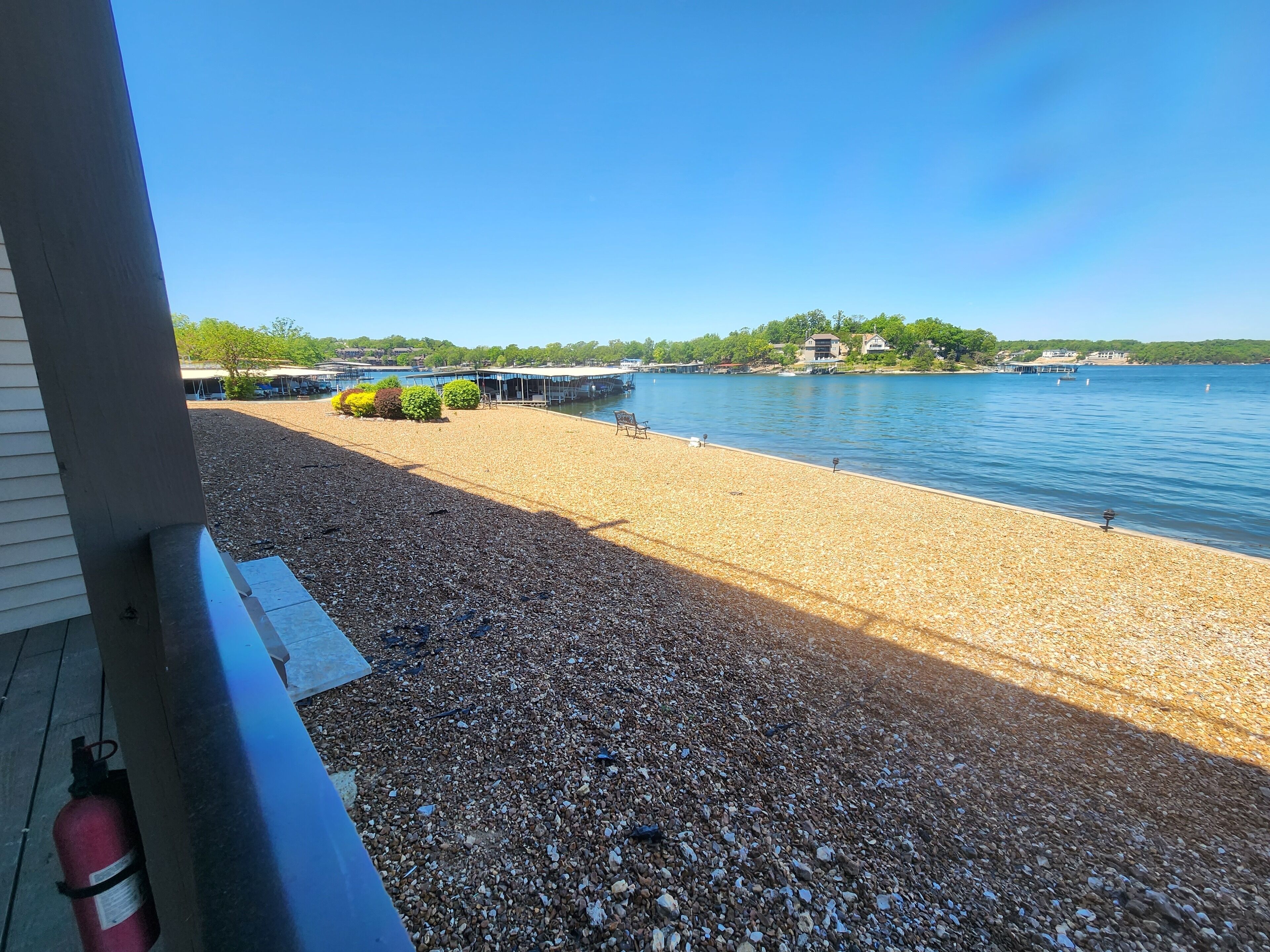 Ein Kiesstrand mit freiem Blick auf einen stillen See, einer Anlegestelle und Gebäuden im Hintergrund.