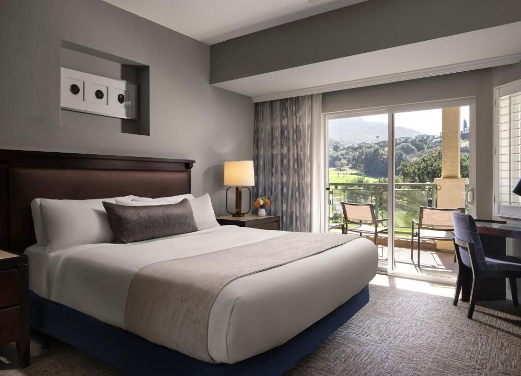 Habitación de hotel con una cama grande, un escritorio con silla, una lámpara y vistas a un paisaje verde a través de una puerta corrediza de vidrio.