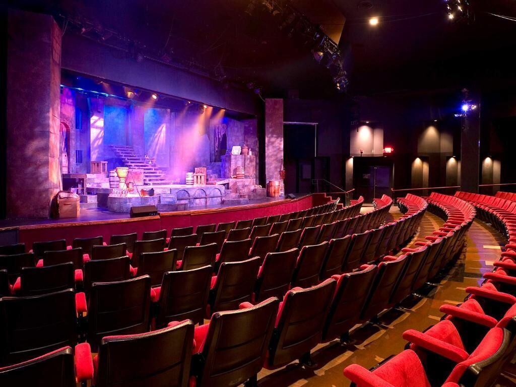 Ein Auditorium mit Reihen roter Sitze, einer Bühne mit blauem Licht und einem Hintergrund, der einer Stadtansicht ähnelt.