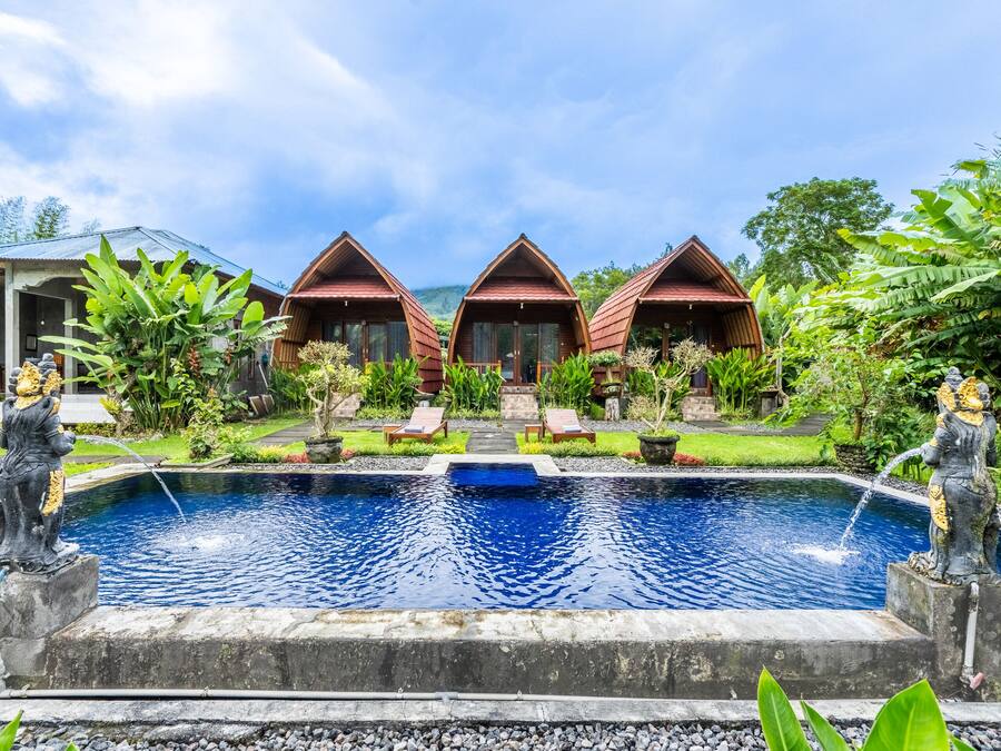 Un resort con tres cabañas de madera, una piscina central con estatuas y abundante vegetación.