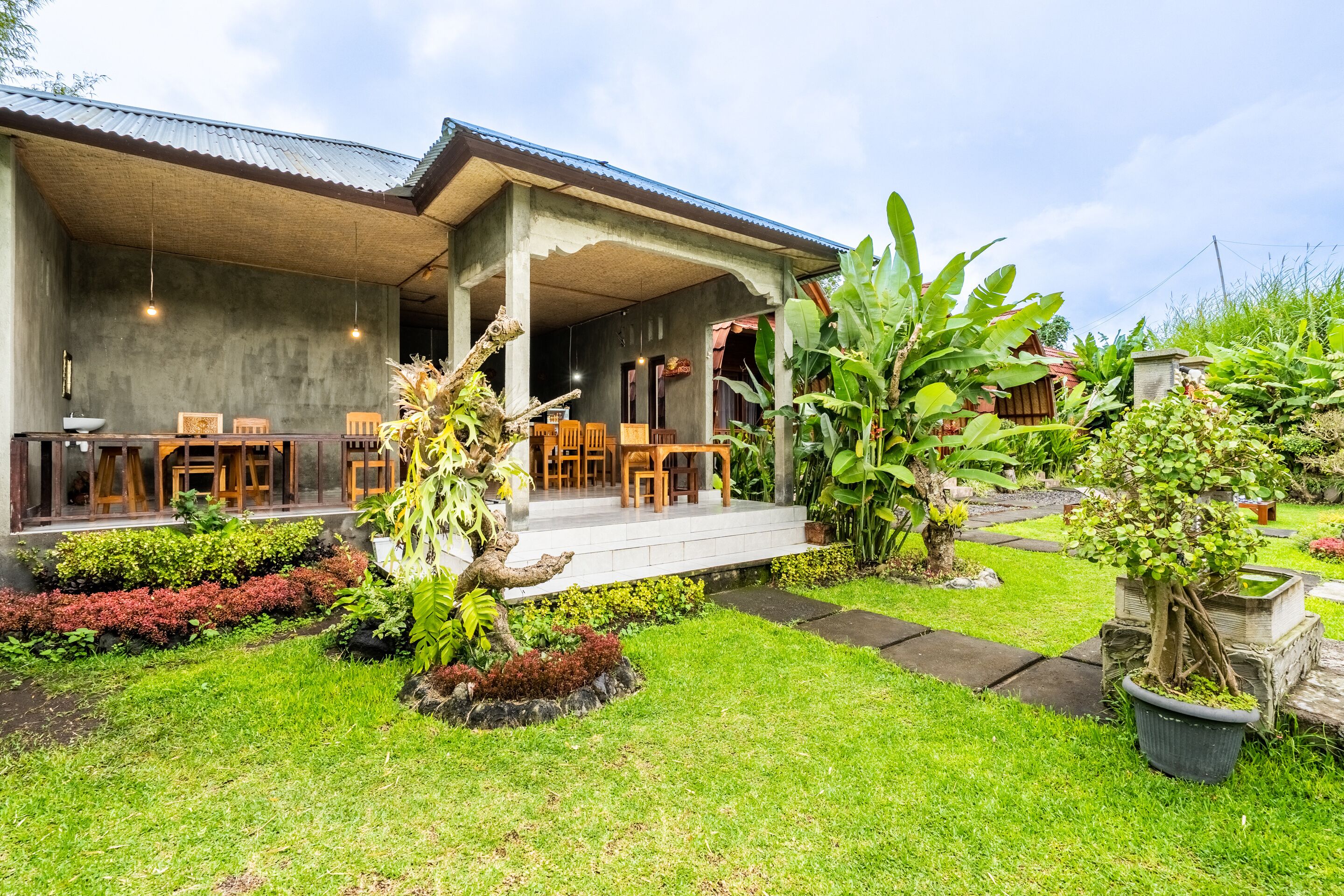 A covered outdoor seating area with tables and chairs, surrounded by lush greenery and potted plants.
