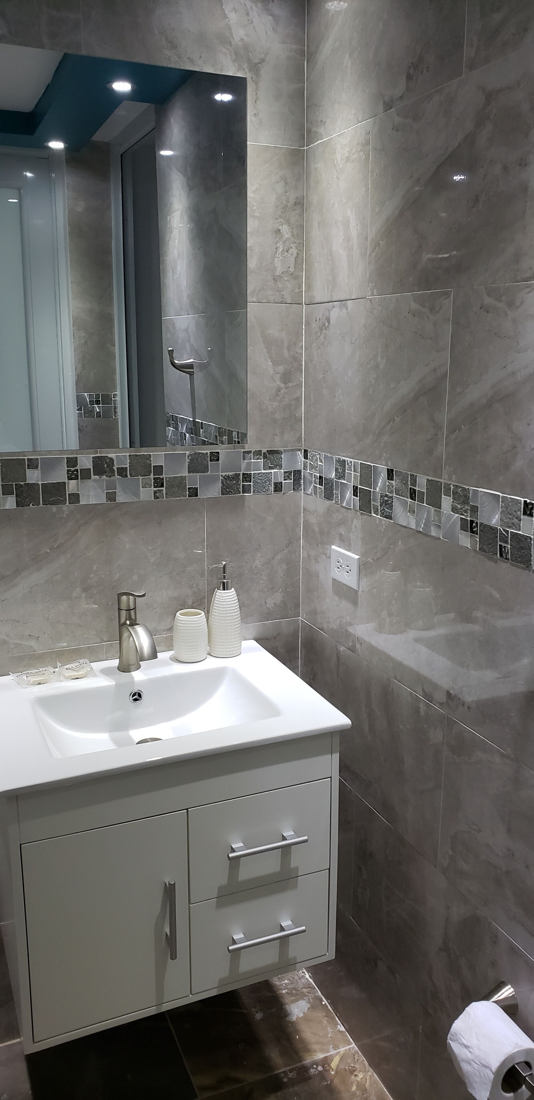 A modern bathroom with a white sink, stainless steel faucet, and a mirror above it. There are two soap dispensers and a small mirror on the sink. The walls are tiled with a gray marble pattern, and there is a tiled backsplash with a mosaic design.