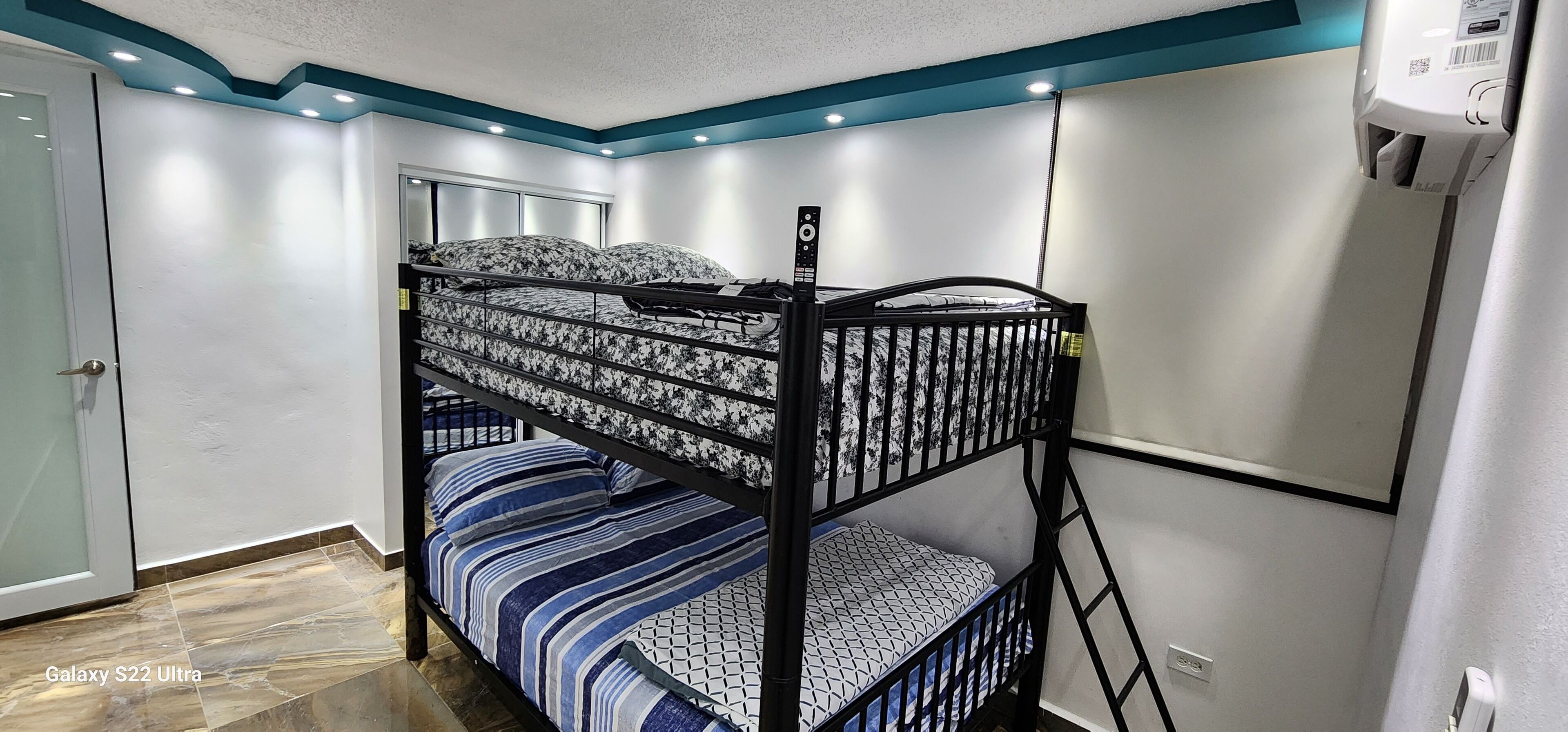 A bunk bed with a patterned mattress, a lower bunk with a striped blanket, and a ceiling with recessed lighting.